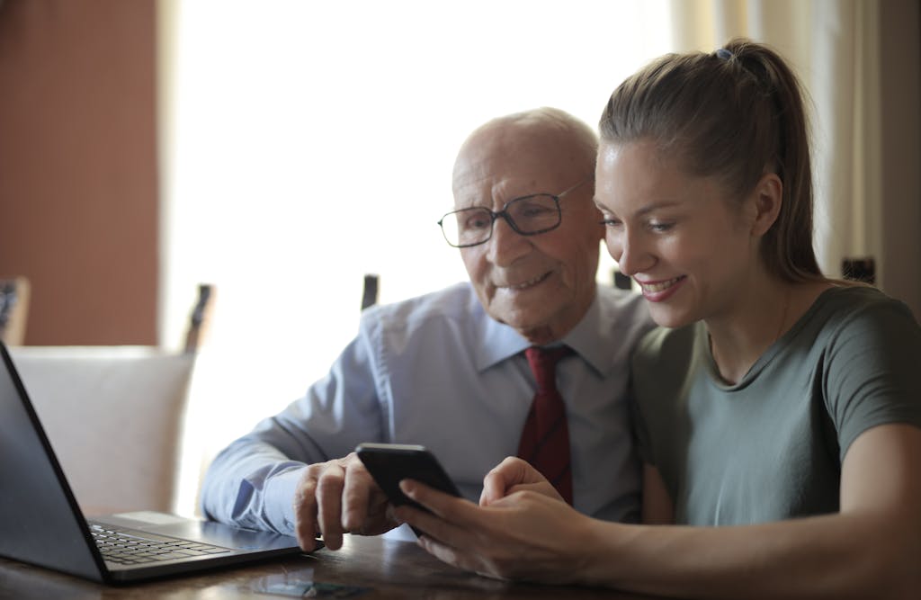 Positive senior man in formal shirt and eyeglasses and smiling young granddaughter sharing mobile phone while sitting near laptop at table